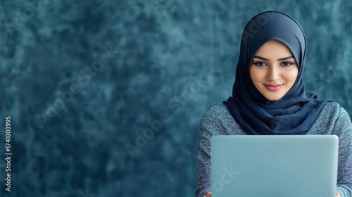 Young Muslim woman wea hijab using laptop computer at home with a calm focused expression and a textured blue wall background for online learning work or communication