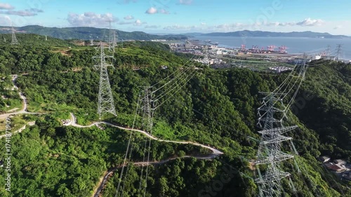 Transmission towers with power lines stand on a lush green hill under a blue sky