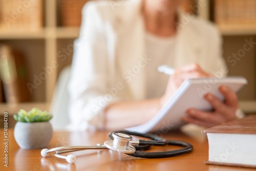 Healthcare professional takes notes during a consultation in a bright doctor's office, stethoscope and medical book on the desk as patient information and prescription details are recorded