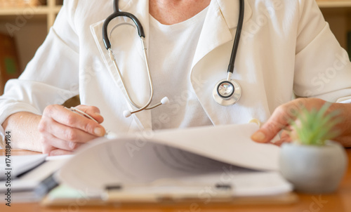 Healthcare professional wearing a stethoscope and white coat, writing notes on patient files and reviewing documents during a general practitioner consultation in a doctor's office