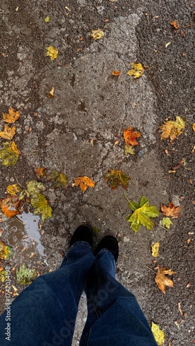 Woman walking in autumn park. A girl in blue wide jeans and black autumn boots stands on rain-wet asphalt surrounded by autumn maple leaves. A top-down, first-person view. Autumn, rainy weather.