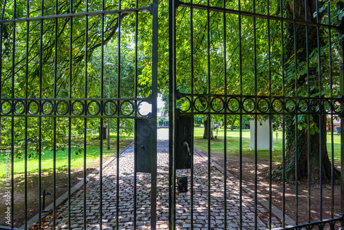 Close-up of bars of half-open black wrought iron gate, entrance to public park, cobblestone path and lush foliage of green trees in background, sunny spring day
