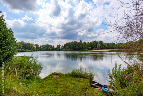 Landscape of leafy trees surrounding lake with mirror reflection on water's surface, green grass on shore, sky with abundant clouds over the blue sky, partly cloudy day in Rekem, Limburg, Belgium