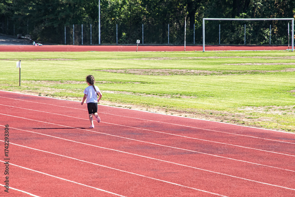 Fototapeta premium A little girl runs alone on the red tartan track of a small stadium during a sunny day