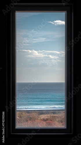 Serene ocean view through a dark framed window, with gentle waves lapping a rugged shore under a vast blue sky.