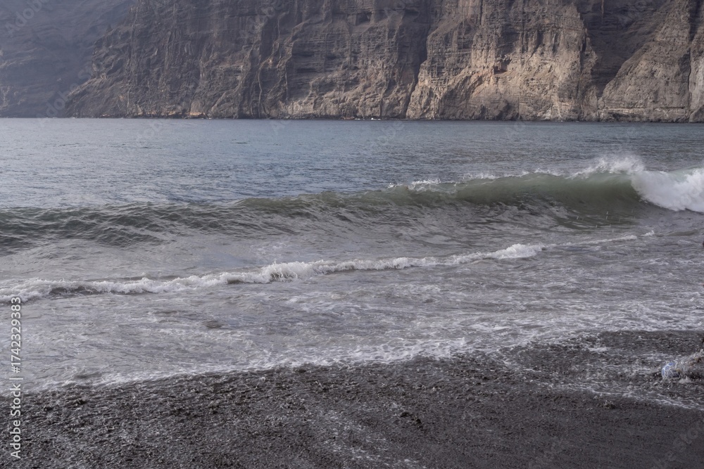 Fototapeta premium Ocean wave rolling on black volcanic sand beach with foamy surf and reflection of cloudy sky near Los Gigantes, Tenerife, Canary Islands, Spain. Beautiful seascape nature background.