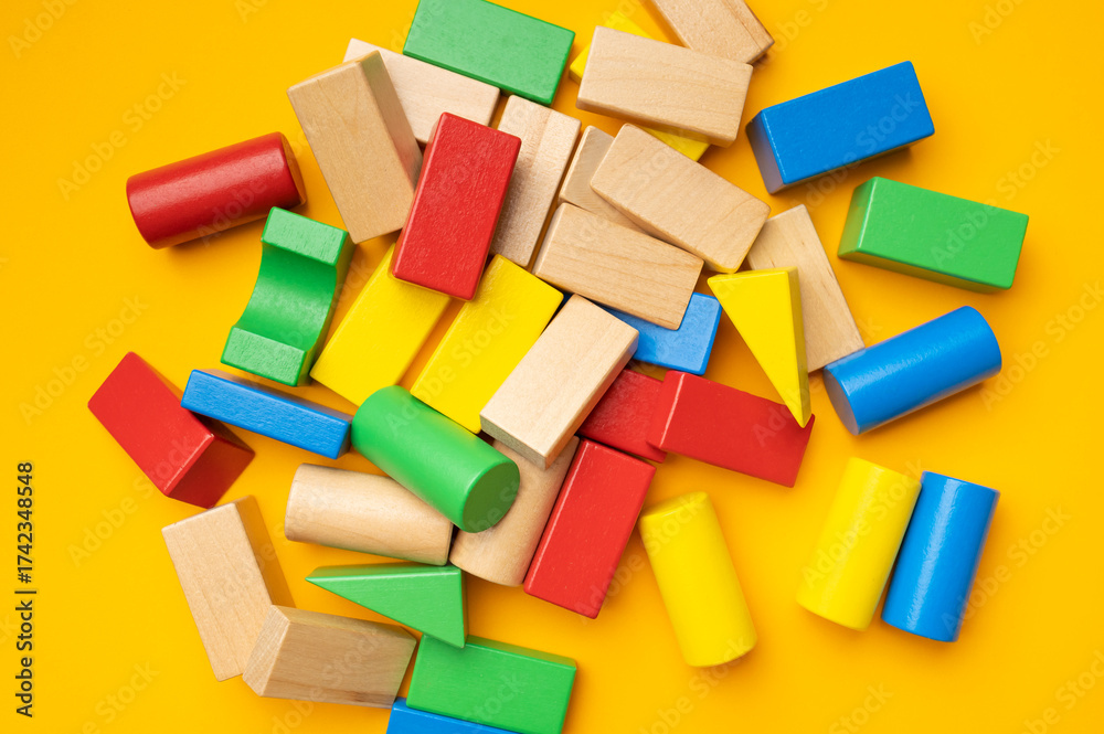 Naklejka premium Wooden children's multi-colored bright construction set blocks of different shapes on yellow background. Montessori style cubes game for toddlers flat lay top view. Concept of learning and education