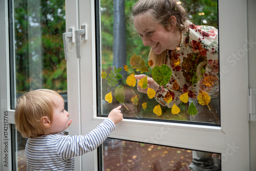 Young mother and her 15-month-old son playing creatively outdoors on a rainy day, decorating the window with colorful autumn leaves, learning and bonding through nature-inspired play