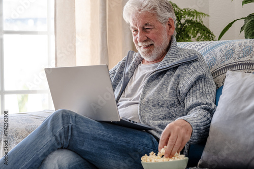 Elderly 70s man seated on sofa browsing on laptop, senior looking at laptop screen surfing the net while eating popcorn, older generation and modern tech application easy usage concept