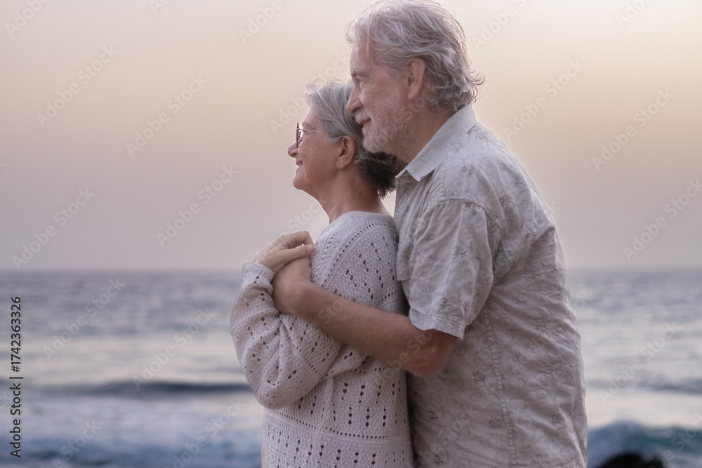 Fototapeta premium Smiling senior couple embraced on the beach in sunset light looking at the horizon over sea - old relaxed elderly couple outdoors enjoying together vacations or retirement