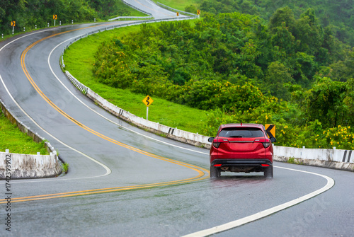 Red car driving on a winding mountain road after rain, scenic travel route surrounded by green hills, concept of road trip, safety, and adventure journey in nature.