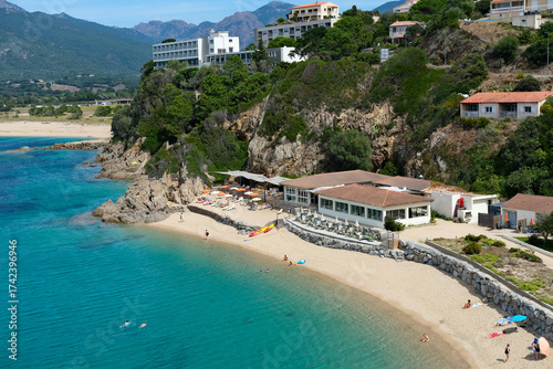 Fototapeta Naklejka Na Ścianę i Meble -  Sandy beach with sunbathers and swimmers, turquoise sea, and a cliffside resort with terraces and umbrellas.
