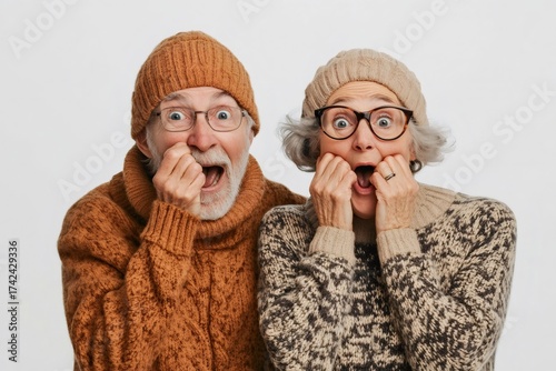 Senior couple widely opening eyes and mouths, holding cheeks, expressing extreme surprise and panic on neutral background