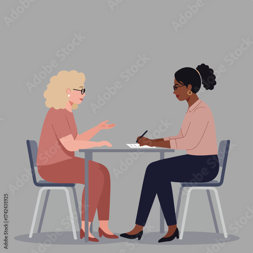 Two women in professional attire sit at a table, one speaking, the other writing