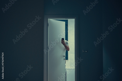 Man partially hidden behind door holding smartphone in dimly lit interior, mysterious atmosphere. Hand with smartphone visible through open door, moody low light interior scene with suspenseful feel.