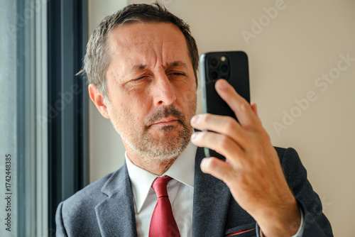Farsighted businessman squinting and struggling to read a text message on smartphone in bright office interior. Mature adult entrepreneur with vision problems trying to read phone screen in workplace.