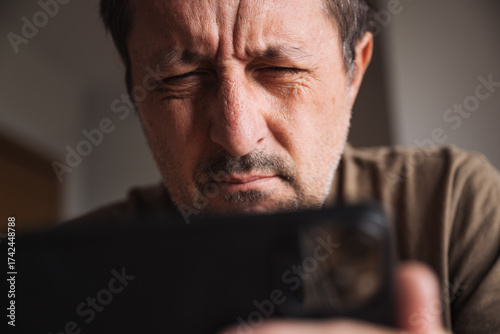 Farsighted mature man squinting eyes while reading message on smartphone screen indoors.