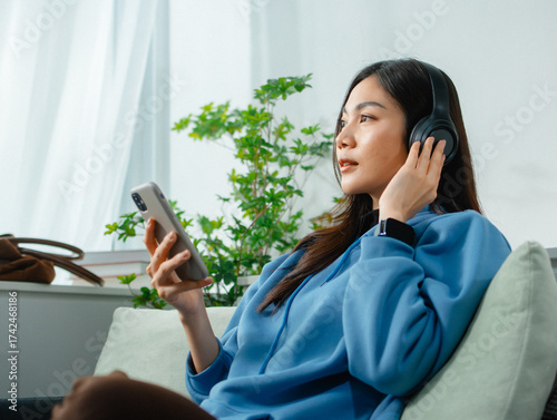 Young Asian woman Relaxing at home wearing headphones Listening to Music Streaming on Mobile Phone app sitting on sofa. Asian female using smartphone listen to podcast, browsing social media at home.