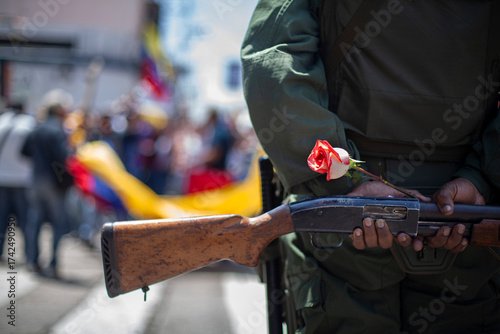 A Venezuelan soldier from behind holds a rose, a symbol of violence and peace, next to his weapon.