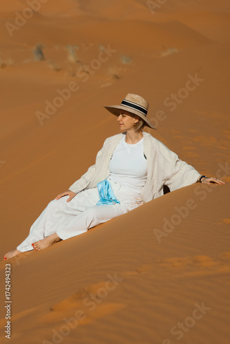 A girl is sitting on a dune in the Sahara Desert, wearing a white dress and a hat.