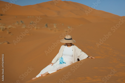 A woman wearing a hat and white clothes is sitting on the dunes of the Sahara Desert