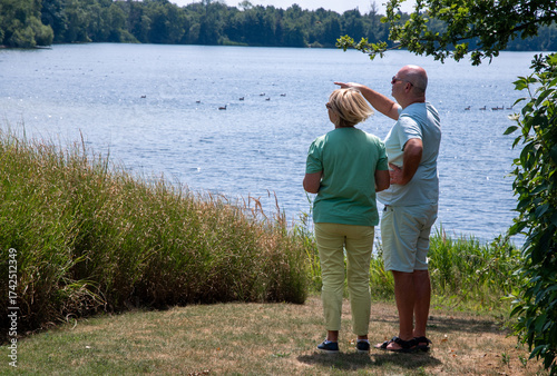 Mature couple standing together by a serene lake. Enjoying the sunny weather and observing ducks swimming. With the man pointing towards the water. Sharing a moment of leisure and connection in nature