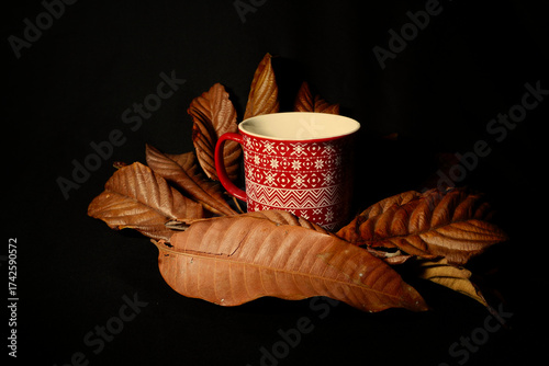 Autumn still life with red cup among dry leaves on dark background. Warm seasonal mood evoking fall evenings and cozy atmosphere, also suitable for Halloween themes.