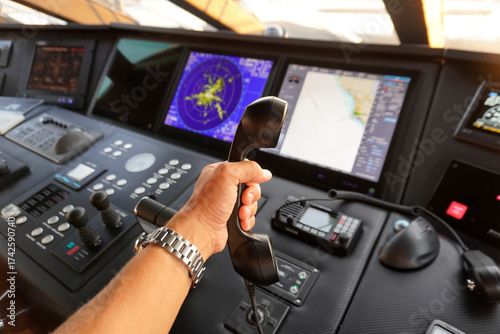 Close-up of a hand holding a marine VHF radio handset on a ship’s bridge with radar and navigation displays. Modern maritime communication and navigation equipment.