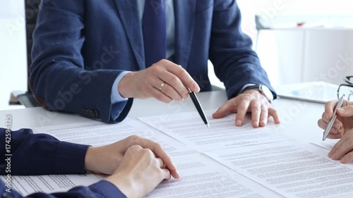 Business professionals gather in a bright office around a desk strewn with legal documents and pens, reviewing paperwork and preparing to sign a formal contract after negotiations