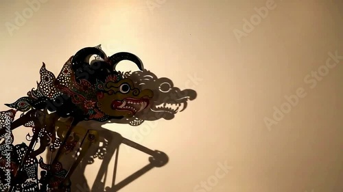 An intricately designed Indonesian Wayang Kulit shadow puppet is displayed with a moving light source altering the shadow it casts on the wall.