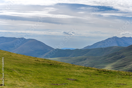 Scenic view of a mountain range with grass and pastures, a small peak in the background, and clouds above on a calm Pyrenean afternoon.”