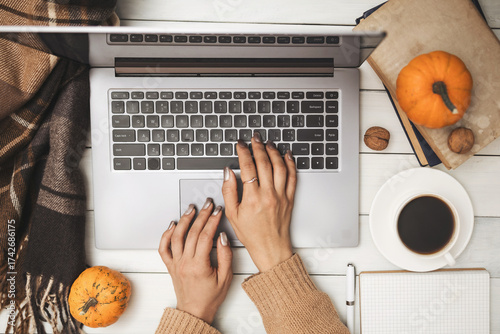 Female hands in sweater typing on laptop on white wooden background with pumpkins, coffee cup and notebook, cozy autumn workspace scene, top view