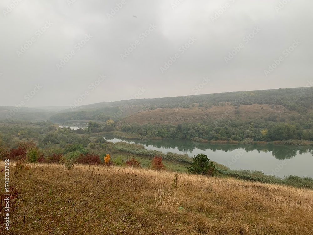 Naklejka premium Misty autumn morning with fog over a calm pond surrounded by colorful trees on gentle hills. Peaceful natural landscape in soft light.