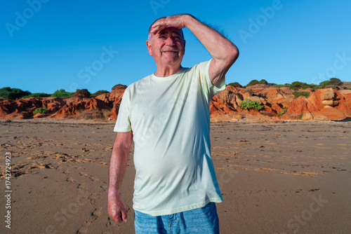 Hombre mayor dándose sombra con la mano para ver mejor, un día soleado en la playa con cielo azul.