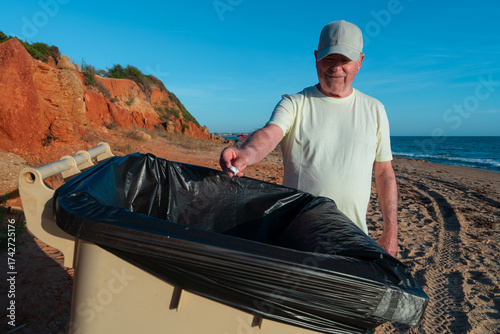 Hombre mayor tirando un papel a la basura en la  playa un día soleado con cielo azul.