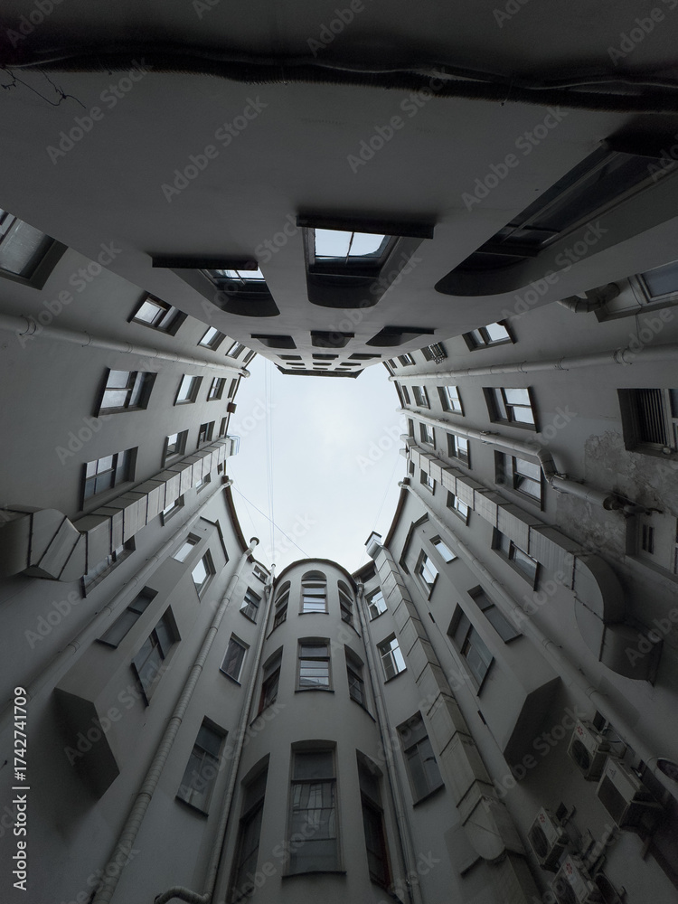 Fototapeta premium Dramatic upward view between tall grey apartment blocks showcasing windows balconies pipes and curved architecture converging toward bright sky