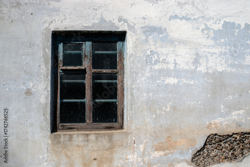 A white wall of an old house with a window. A window on the wall of an old building. The sun illuminates the wall where the plaster and exterior coating have peeled.