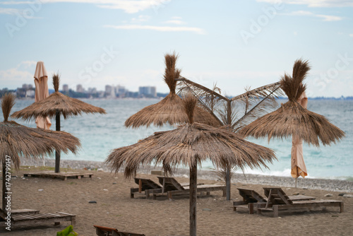 The shoreline of a sea beach is made of small pebbles. On the beach are sun umbrellas under a palm-tree canopy and sun loungers. In the background is a city.