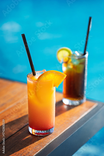 Tequila Sunrise and a Cuba Libre cold cocktails with citrus slices on a wooden table near a blue pool.
