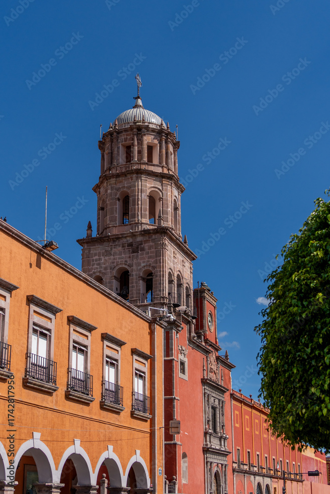 Fototapeta premium A tall tower with a dome sits in front of a row of buildings. The buildings are orange and red. Historic center of Queretaro, colonial architecture, decorations for the celebration of Mexico's Indepen