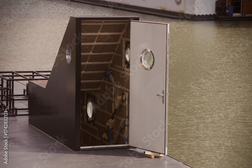 Puerta abierta de un barco en un canal, con interior sin terminar, ojos de buey y estructura de madera. Detalle arquitectónico junto al agua. Roterdam