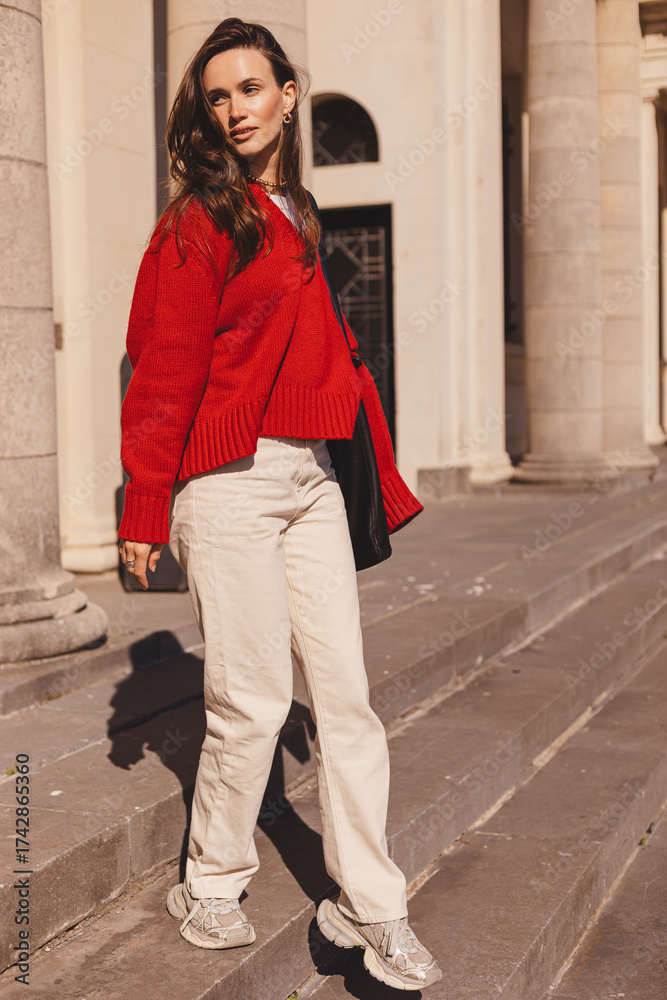 Fototapeta premium Elegance long hair brunette woman go down stairs, wear red sweater, beige pants, she go out from university or museum, old building. Smiling woman turn her head back. Full length portrait.