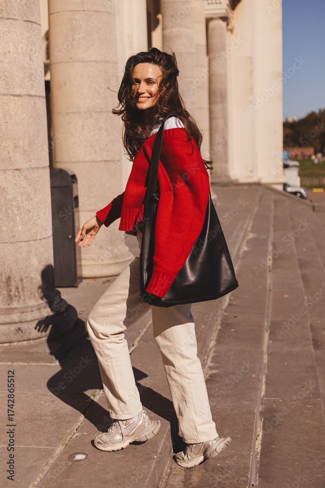 Naklejka premium Photo of lovely positive cute long hair brunette woman going upstairs in old building, she look happy, wear red sweater, beige pants and black bag style fashion outfit. Full length portrait. 