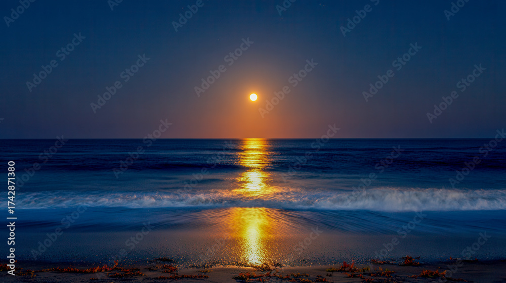 Obraz premium Full moon glowing brilliantly over calm dark ocean waters reflecting golden light onto gentle waves approaching sandy shore at night under clear sky