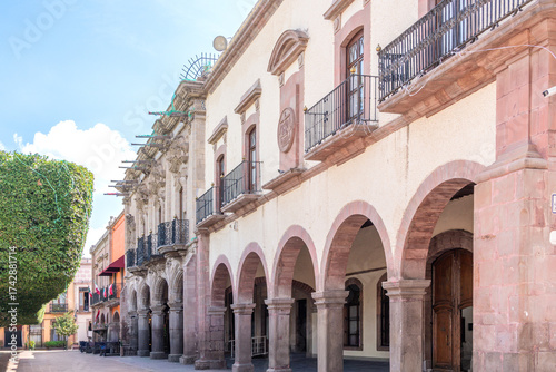 A street with a row of buildings, one of which has a balcony. The buildings are old. Historic center of Queretaro, colonial architecture, decorations for the celebration of Mexico's Independence Day.