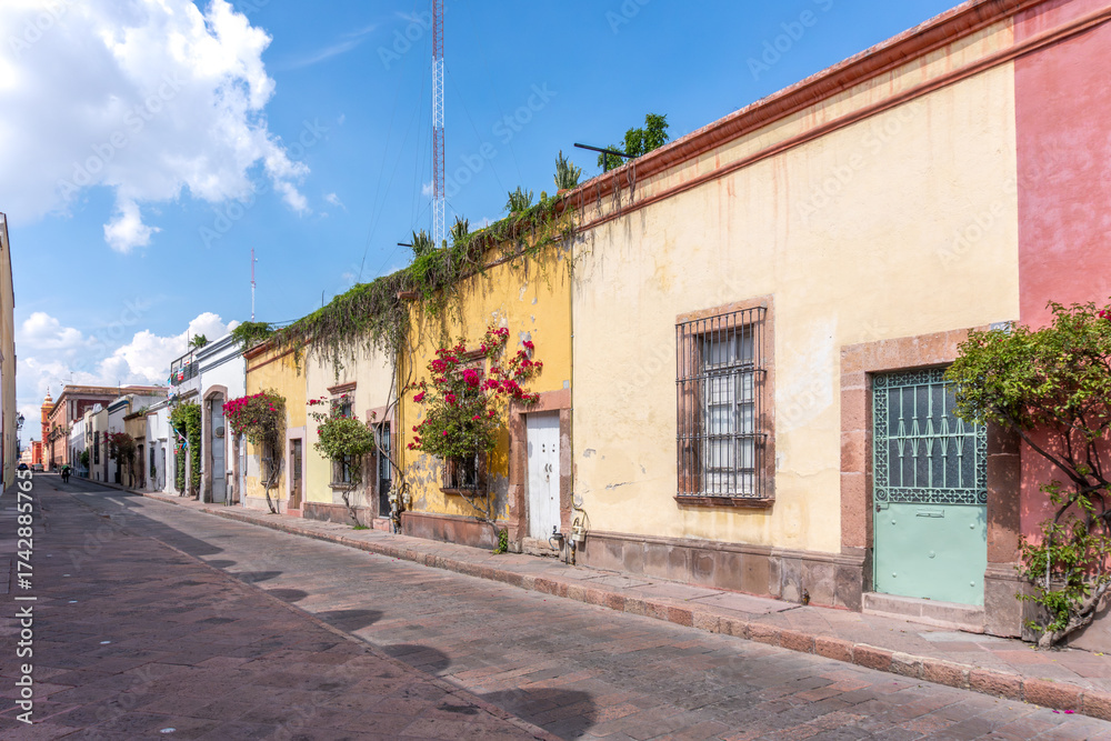 Naklejka premium A street with a row of yellow buildings and a green door. The street is empty and the sky is blue. Historic center of Queretaro, colonial architecture, decorations for the celebration of Mexico's Inde
