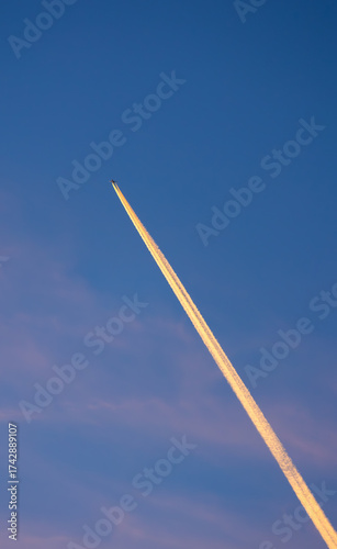 Jet plane flying far away with long diagonal pink contrail against blue sky at sunset, reach for the stars, travel, ambition, direction, farewell