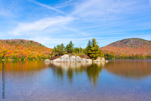 Island framed by two mountains in full autumn colors, at Lake Stukely, Mont-Orford National Park, Quebec, Canada. Full sunshine and rich blue sky with wispy clouds.