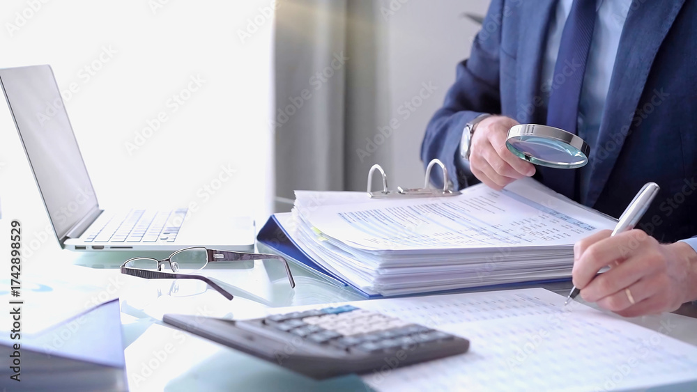 Obraz premium Businessman using a calculator and magnifying glass while analyzing financial documents at desk in fair modern office. Audit and taxes in business