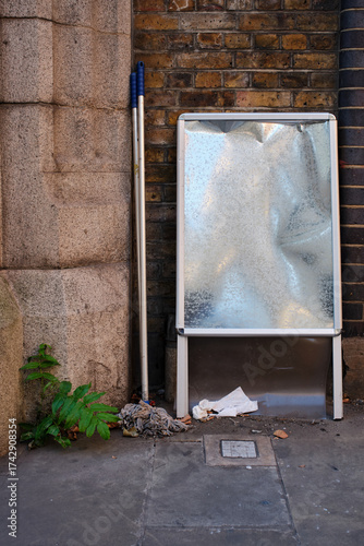 Empty metal sign pavement sign and two mops against a brick wall with some rubbish garbage litter